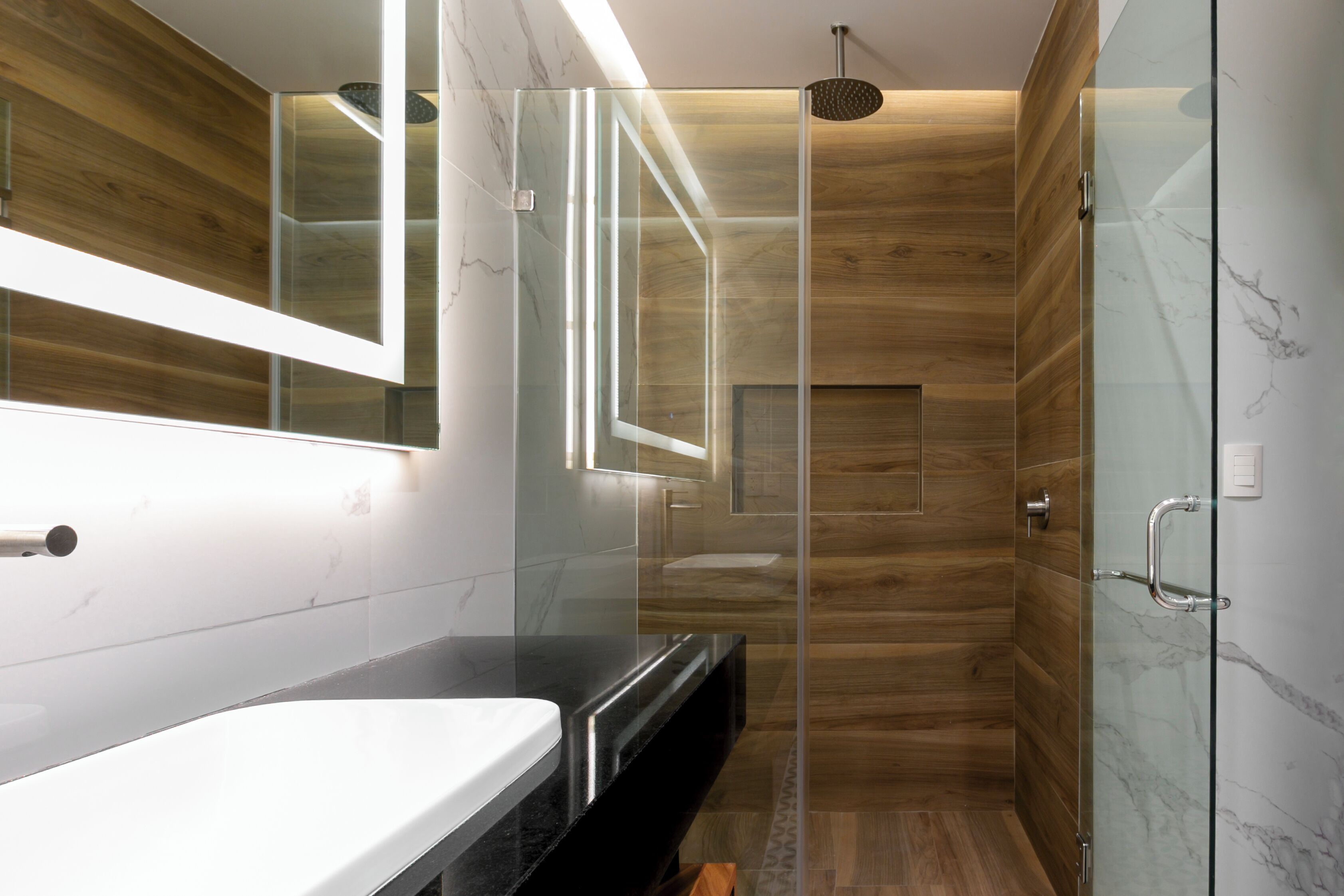 Un baño moderno con ducha envuelta en vidrio, tocador de madera y salpicadero de mármol.
