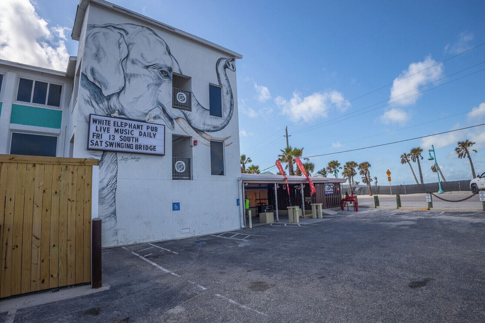 A building with a mural of an elephant, a sign for the White Elephant Pub, and a parking area with palm trees in the background.