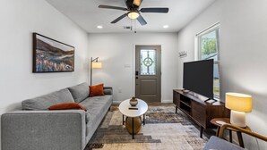 A modern living room with a grey sofa, a coffee table, a TV, and a ceiling fan. - Cozy & Bright 2BR Home - 10 Min to Downtown (Houston)