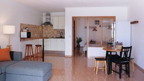 Un saló modern amb un sofà gris, tamborets de fusta, una taula de dinar amb caires i una cuina amb armaris blancs i un microones.