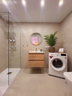 A modern bathroom with a glass shower enclosure, a built-in sink with a mirror, a wall-mounted washing machine, and a potted plant.
