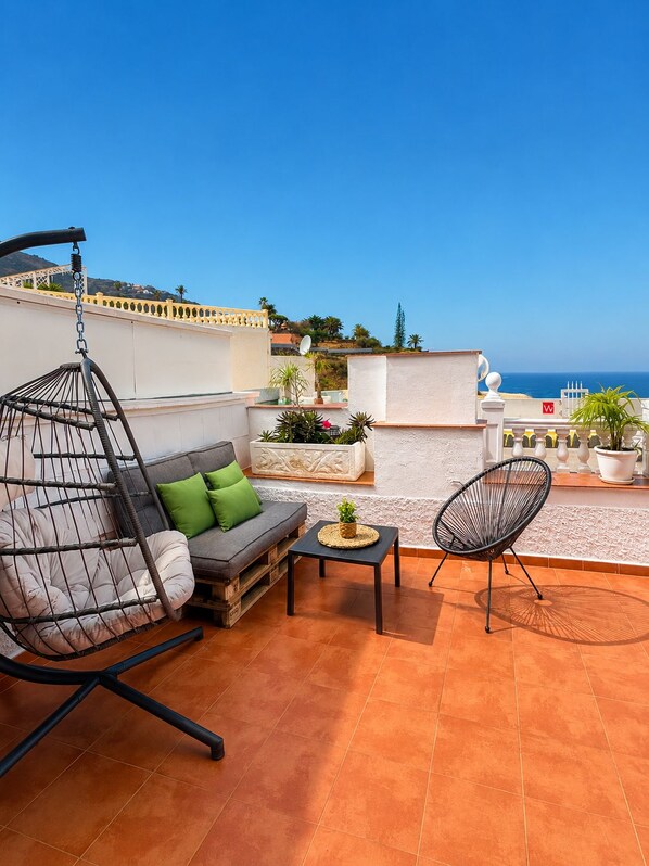 A rooftop terrace with a hanging chair, a sofa with cushions, a small table, and a chair.