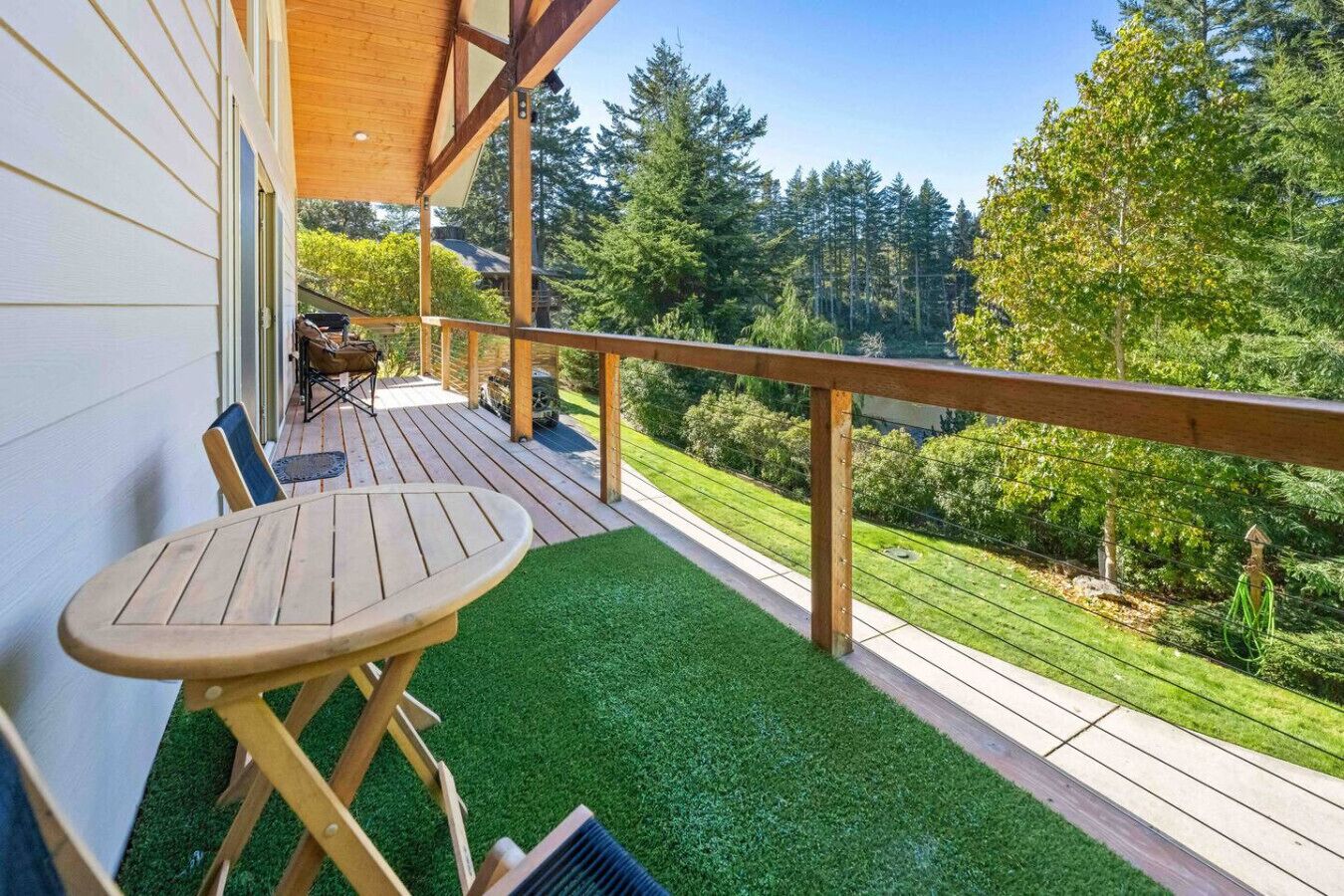 A wooden deck with a table and chairs, surrounded by green artificial grass and a view of a forested area.