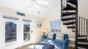 Un salon avec un canapé bleu, un tapis à motifs, un ventilateur de plafond, un escalier en colimaçon noir et une vue sur l’extérieur.