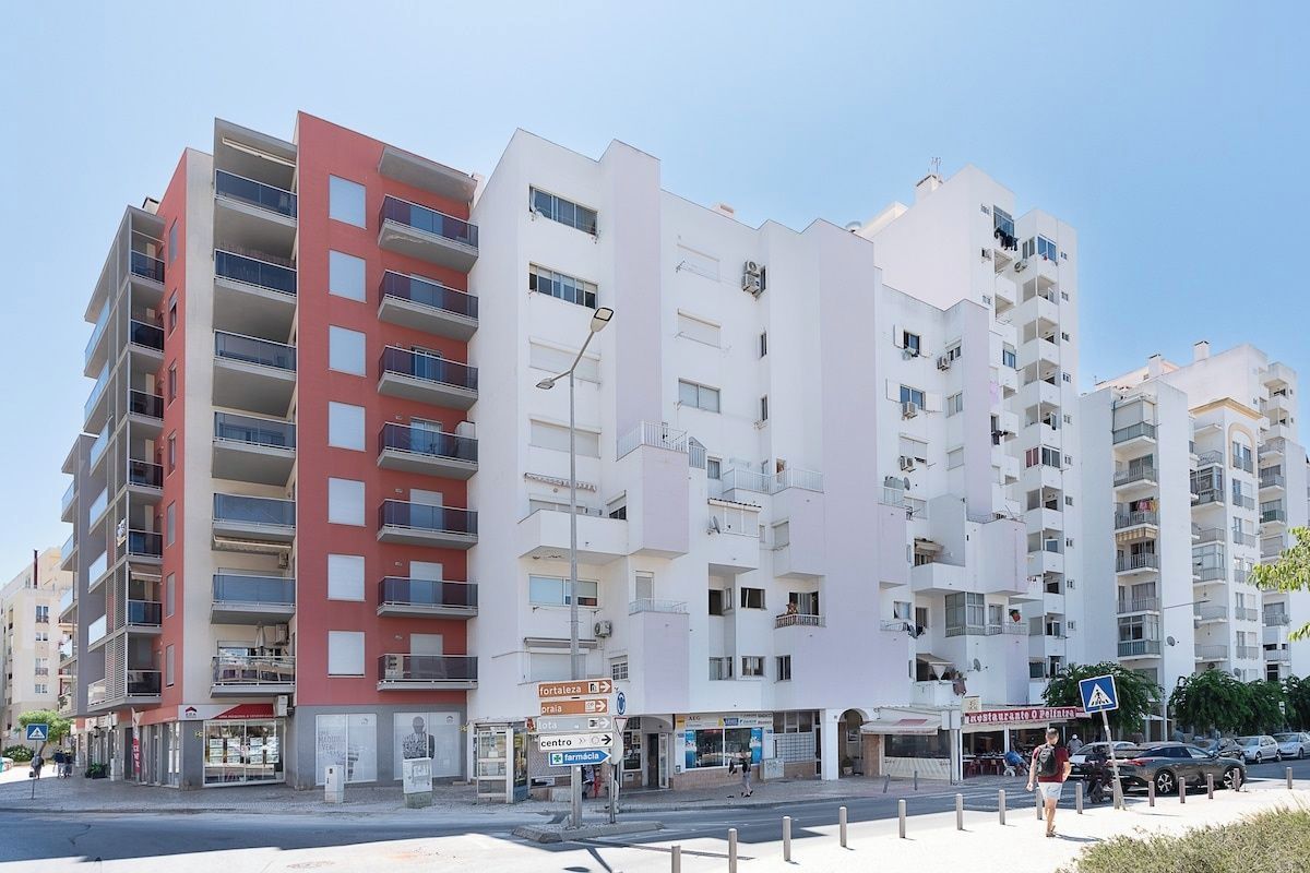 A multi-story residential building with balconies, a street in front, and a clear sky.