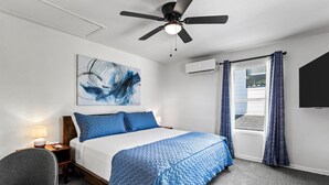 Et hotelværelse med seng, ventilator i loftet, vægmonteret aircondition, et vindue med gardiner, et billede, et sengebord med lampe og en stol.