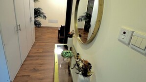 A hallway with a wooden floor, a white wall, a mirror, a small table with a vase of flowers, and a water heater on the wall. - Happy Hostel Dubai (Dubai)