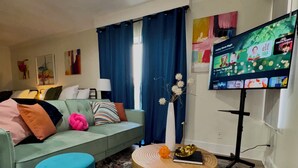 Un salon comprenant un canapé, une table basse ronde, un pouf bleu, un téléviseur posé sur un meuble et un vase orné de motifs floraux.