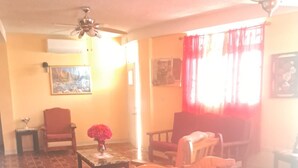 Una sala de estar con un sofá, sillas, una mesa de centro y una mesa de comedor con individuales. Hay una araña, un ventilador de techo y un cuadro en la pared.