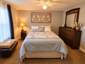 A bedroom with a bed, a nightstand with a lamp, a dresser, a mirror, and a decorative wall piece.