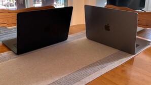 Dos portátiles sobre una mesa de madera con un posavasos tejido, una ventana que muestra muebles de exterior, y un televisor en la pared.