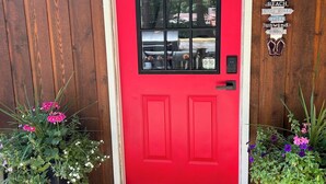 Una puerta roja con un ventanal negro, flanqueada por dos macetas con flores rosadas.