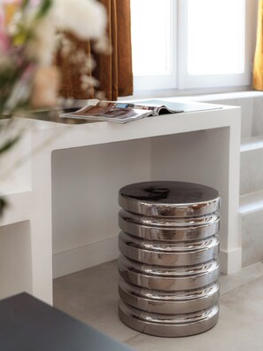 A modern, white, multi-tiered metallic trash bin in a room with a white console table and a magazine on it.