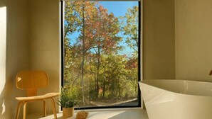 Ein Badezimmer mit Badewanne, einem Holzstuhl, einer Topfpflanze und Blick auf herbstliche Bäume durch ein großes Fenster.