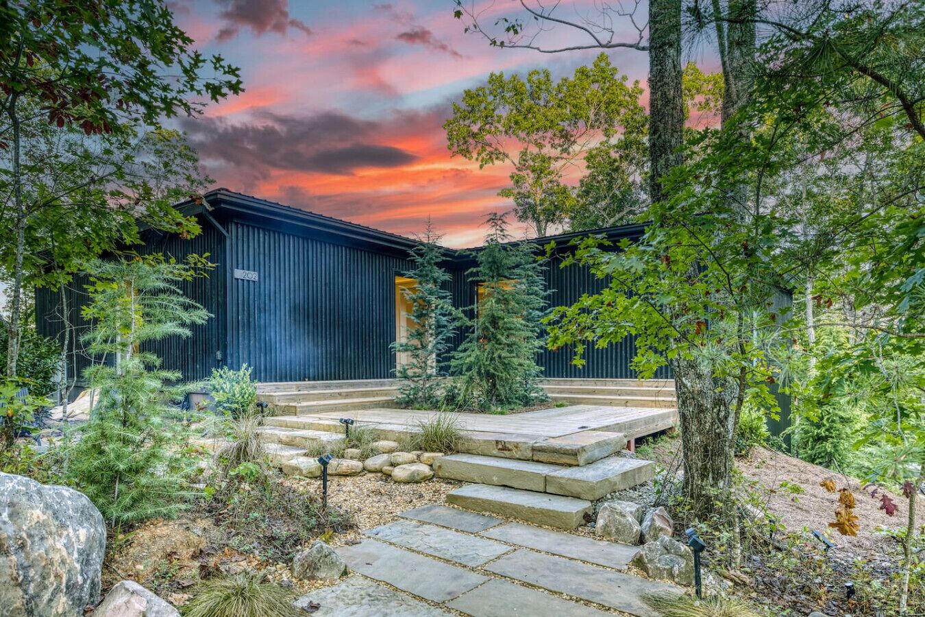 A rustic cabin with a stone staircase leading to a wooden door, surrounded by trees and a sunset sky.