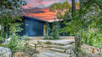 A rustic cabin with a stone staircase leading to a wooden door, surrounded by trees and a sunset sky.
