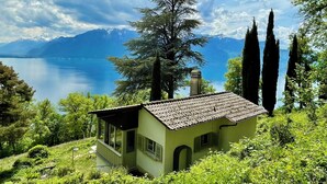 Une petite maison avec un toit de tuiles, entourée d’une végétation luxuriante et de grands cyprès, qui offre une vue sur un lac paisible et des montagnes lointaines.