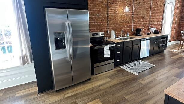 A modern kitchen with a stainless steel refrigerator, black cabinets, a brick wall, and wooden flooring. - Iuka Downtown Historic King Loft B (Iuka)