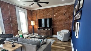 A living room with a brick wall, a ceiling fan, a flat-screen TV, a grey sofa, a coffee table, and a white armchair. - Iuka Downtown Historic King Loft B (Iuka)