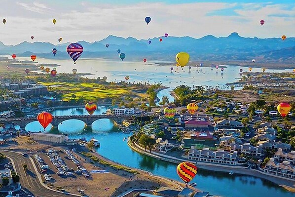 A river with multiple hot air balloons, a bridge, and buildings along the riverbanks.