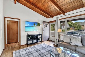 A living room with a wooden ceiling, a large TV on the wall, a sofa, a coffee table, and a view of the outside. - 2BR Cozy Cabin Vibes | King Bed + Vaulted Ceilings (Cheyenne)