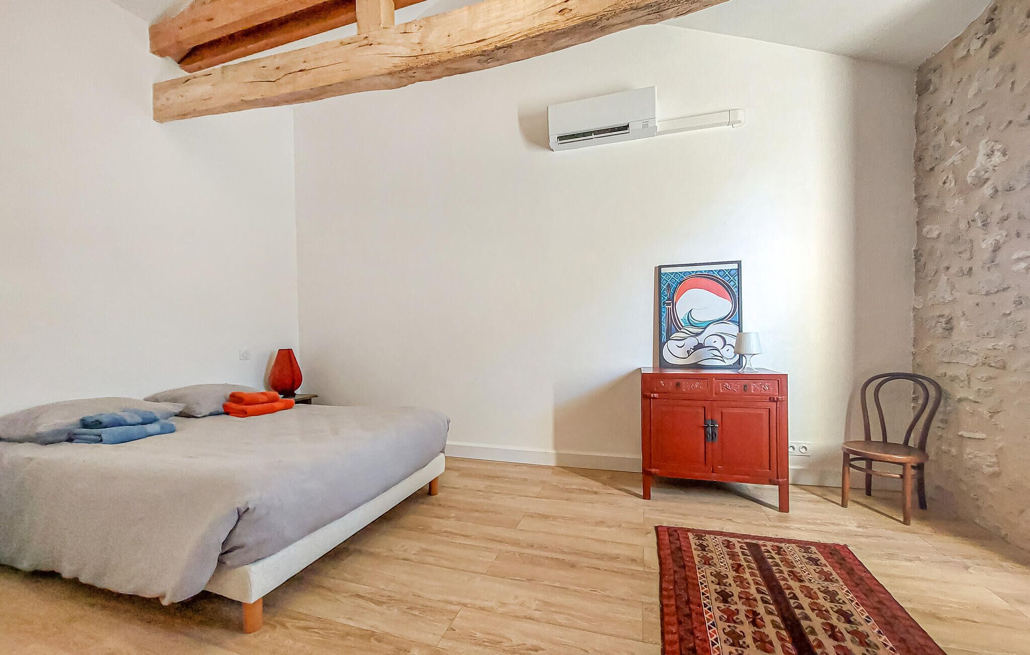 A bedroom with a bed, a red cabinet, a chair, a rug, and an air conditioning unit.
