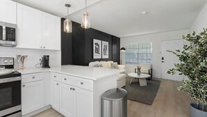 A modern kitchen with white cabinetry, a built-in microwave, and a stainless steel stove. There is a dining area with a white sofa and a small table. The room has a black accent wall with artwork and a large window with blinds. - Luxury apt w/ FREE parking, crib, prime location (Tampa)
