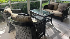 A sunlit patio with wicker furniture, a glass-top table, and a view of greenery through large windows. - 3 Bedroom Condo in Pelican Marsh-Naples, Florida-available for Season 2025-2026. (Naples)