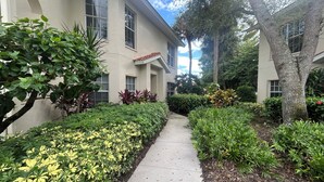 A pathway leading to a building with a beige facade, surrounded by well-maintained hedges and trees. - 3 Bedroom Condo in Pelican Marsh-Naples, Florida-available for Season 2025-2026. (Naples)