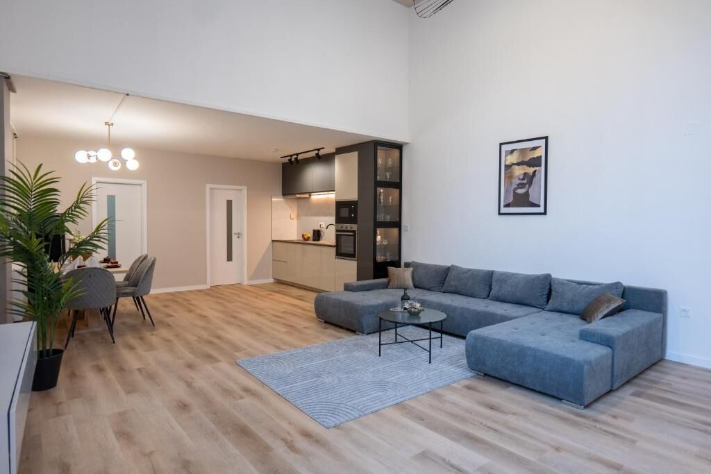 A modern living room with a grey sectional sofa, a dining area with a table and chairs, and a kitchen in the background.