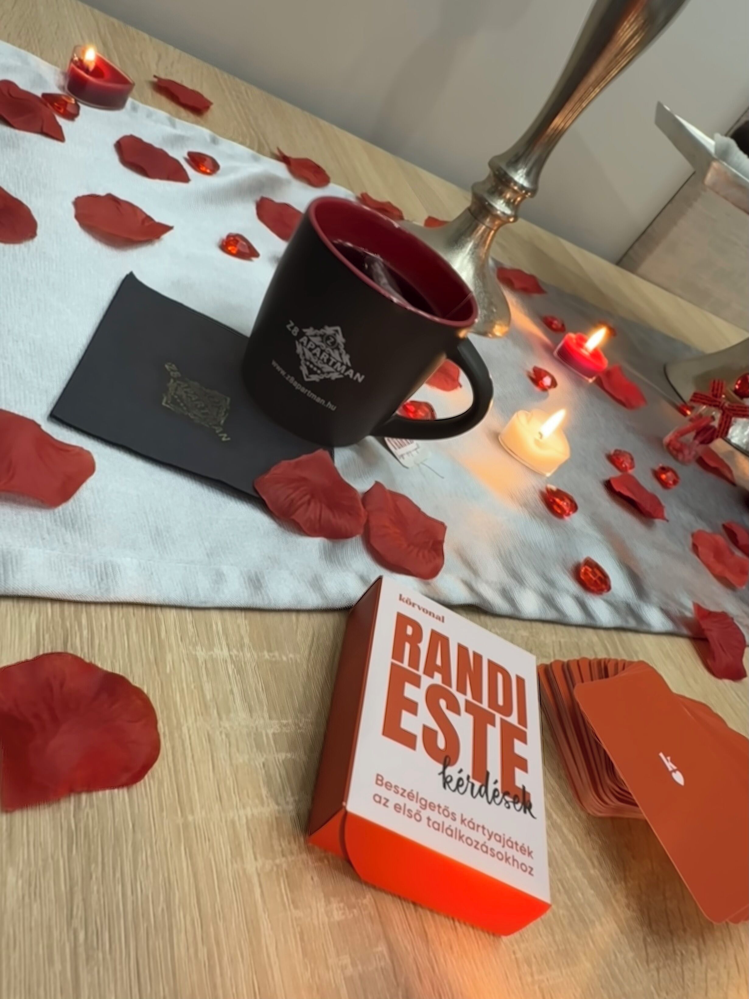 A table with a book titled 'RANDI ESTE' and a black mug with a logo, surrounded by red rose petals and candles.