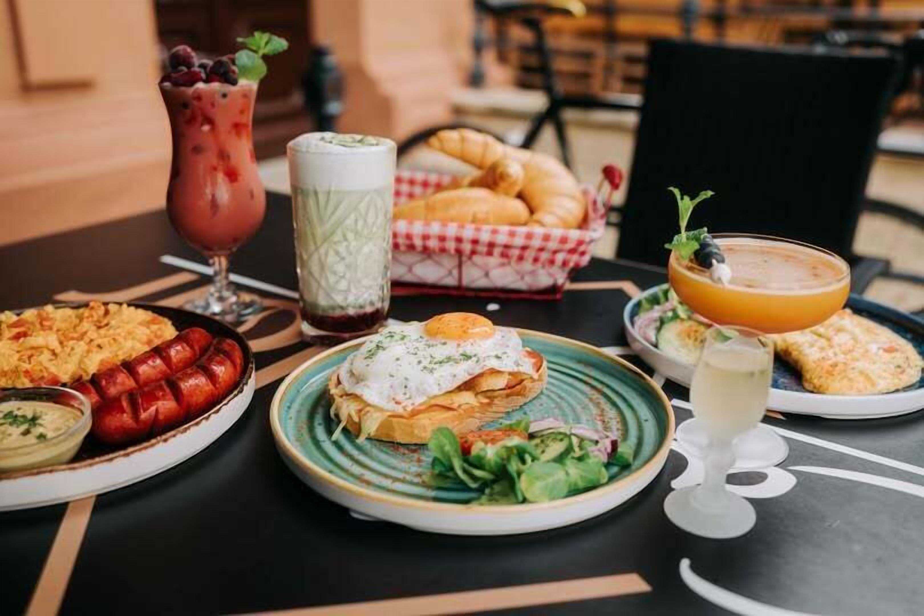 Uma mesa com diversos pratos, incluindo um pão com ovo frito, uma cesta de croissants e um copo de suco de laranja.