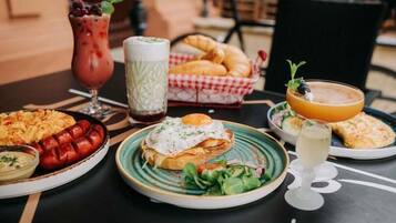 Uma mesa com diversos pratos, incluindo um pão com ovo frito, uma cesta de croissants e um copo de suco de laranja.