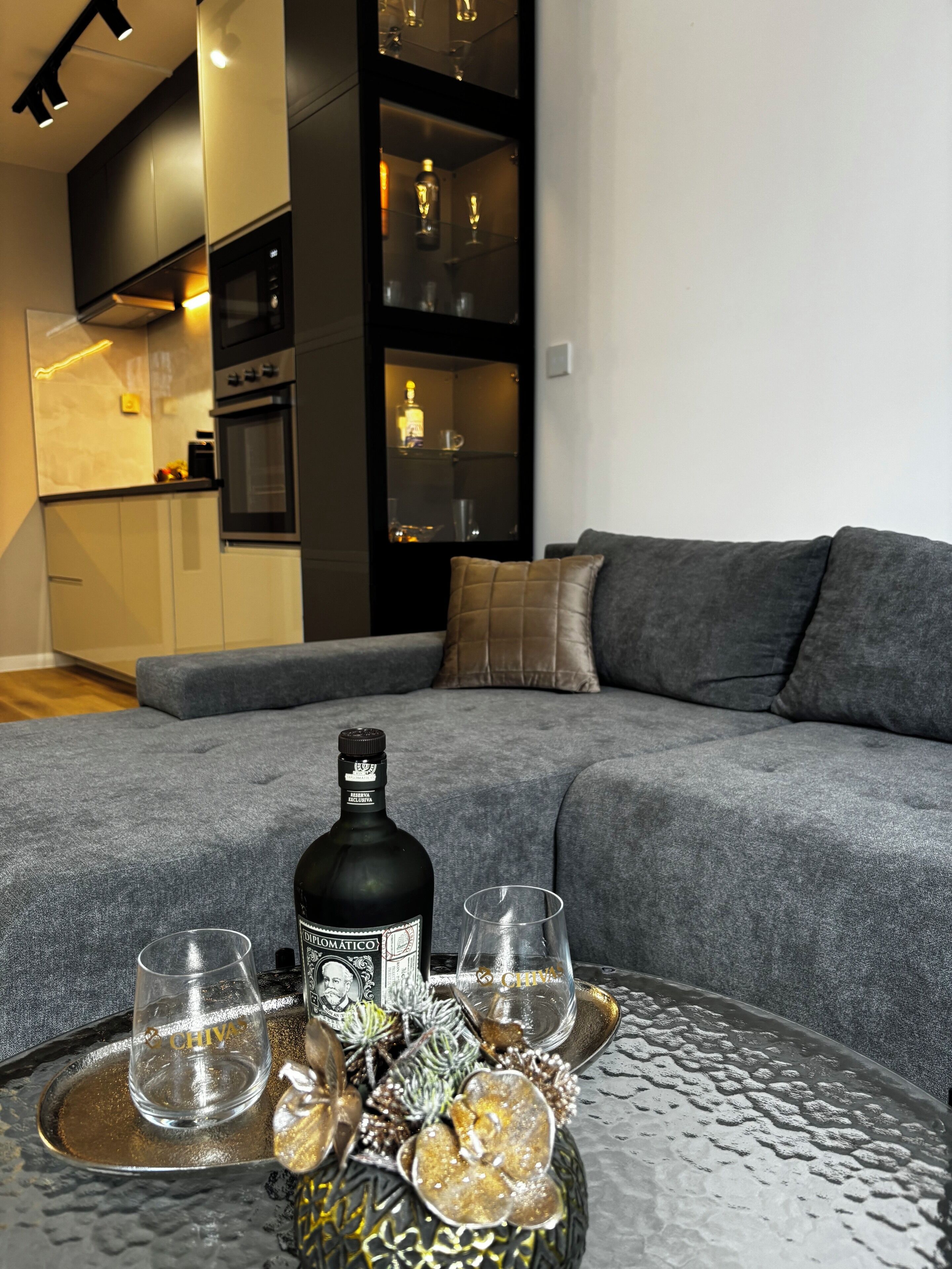 A modern living room with a grey sofa, a dining table with a bottle and glasses, and a kitchen in the background.