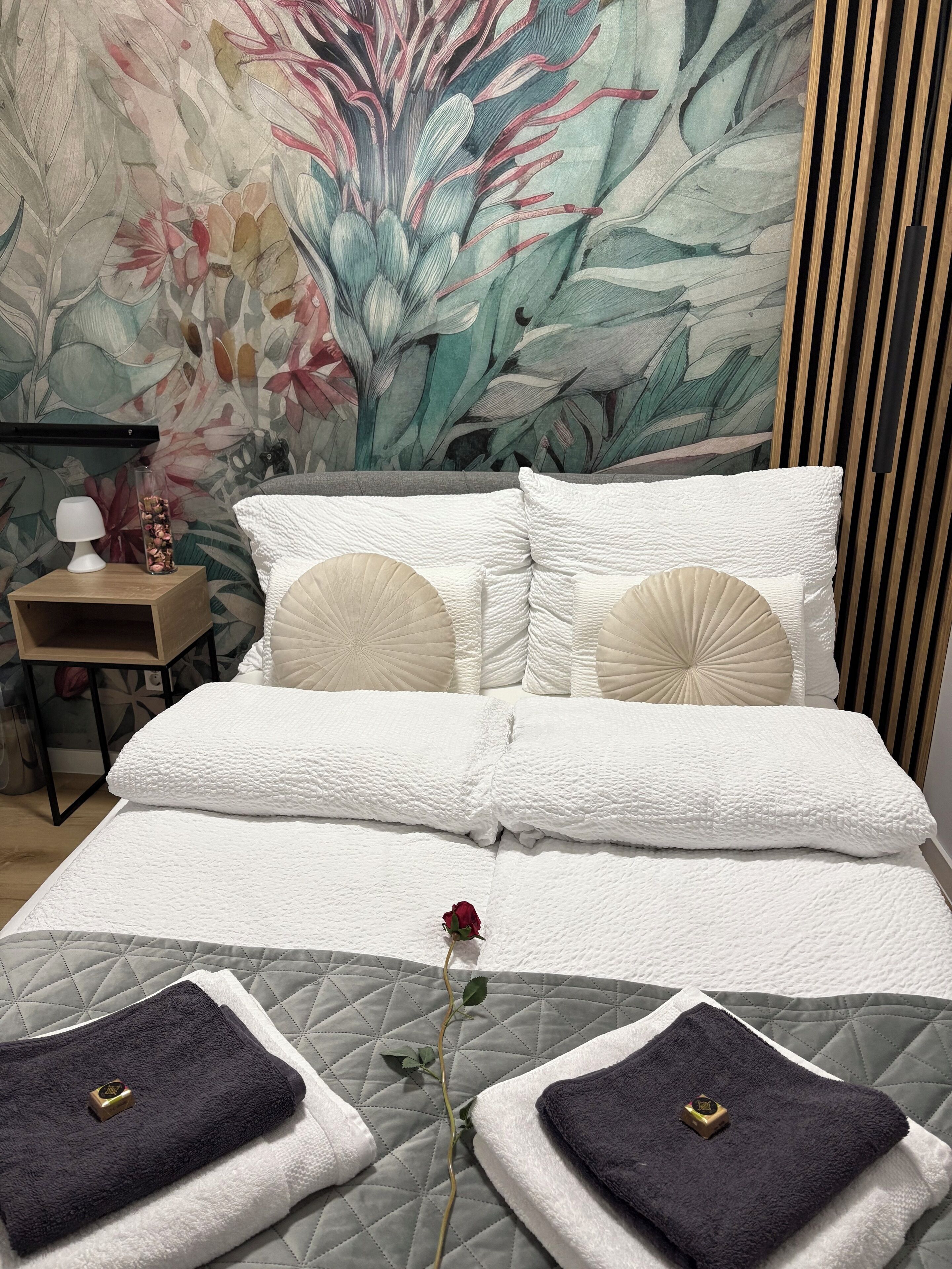 Una cama bien tendida con ropa blanca, un mural decorativo en la pared y una mesita de noche con lámpara y florero.