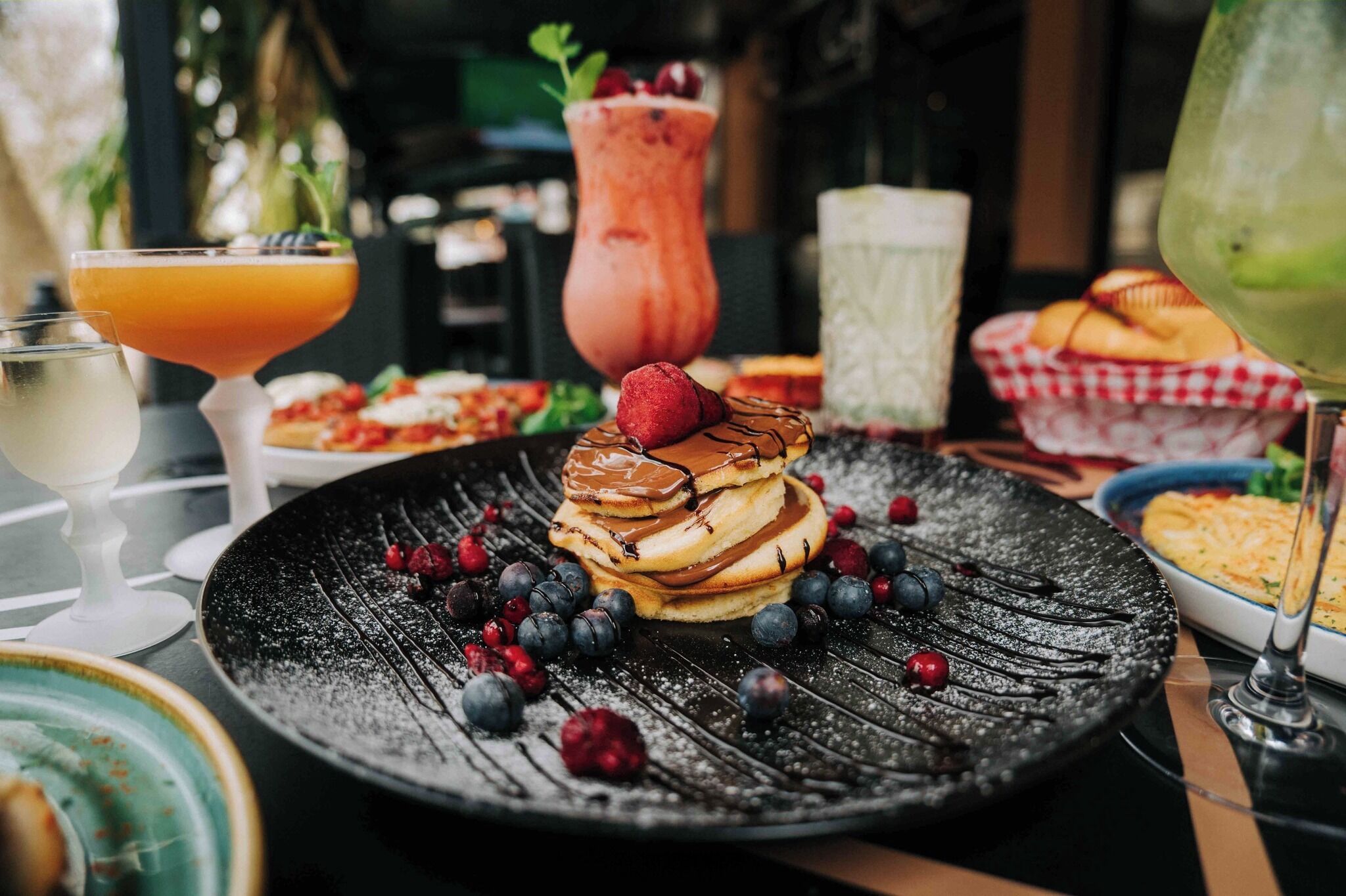 A dining table with a black plate of pancakes topped with strawberries and blueberries, surrounded by various drinks and dishes.