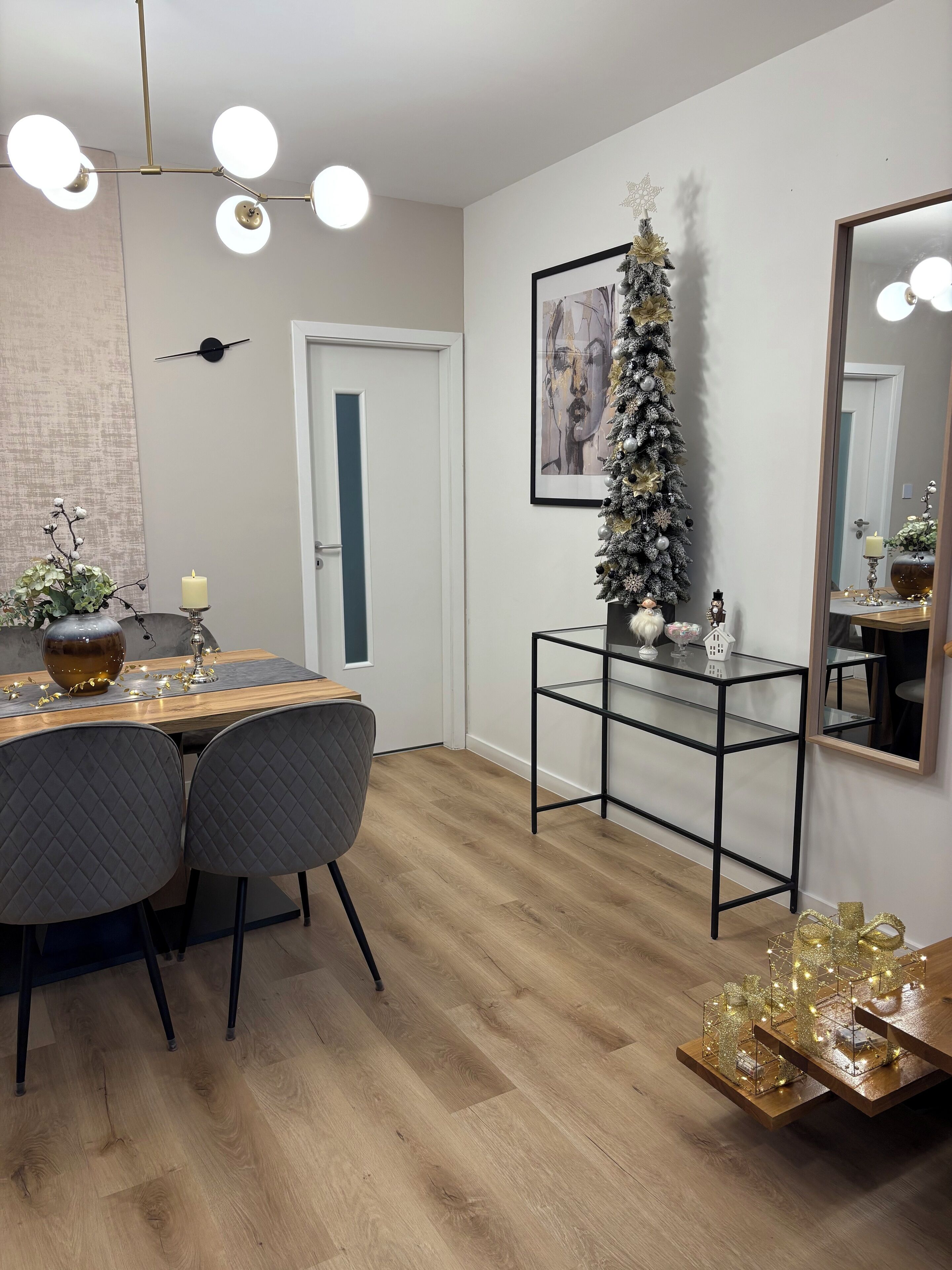 Uma área de jantar moderna com mesa de madeira, duas cadeiras, um pequeno pinheiro de Natal e um espelho decorativo.