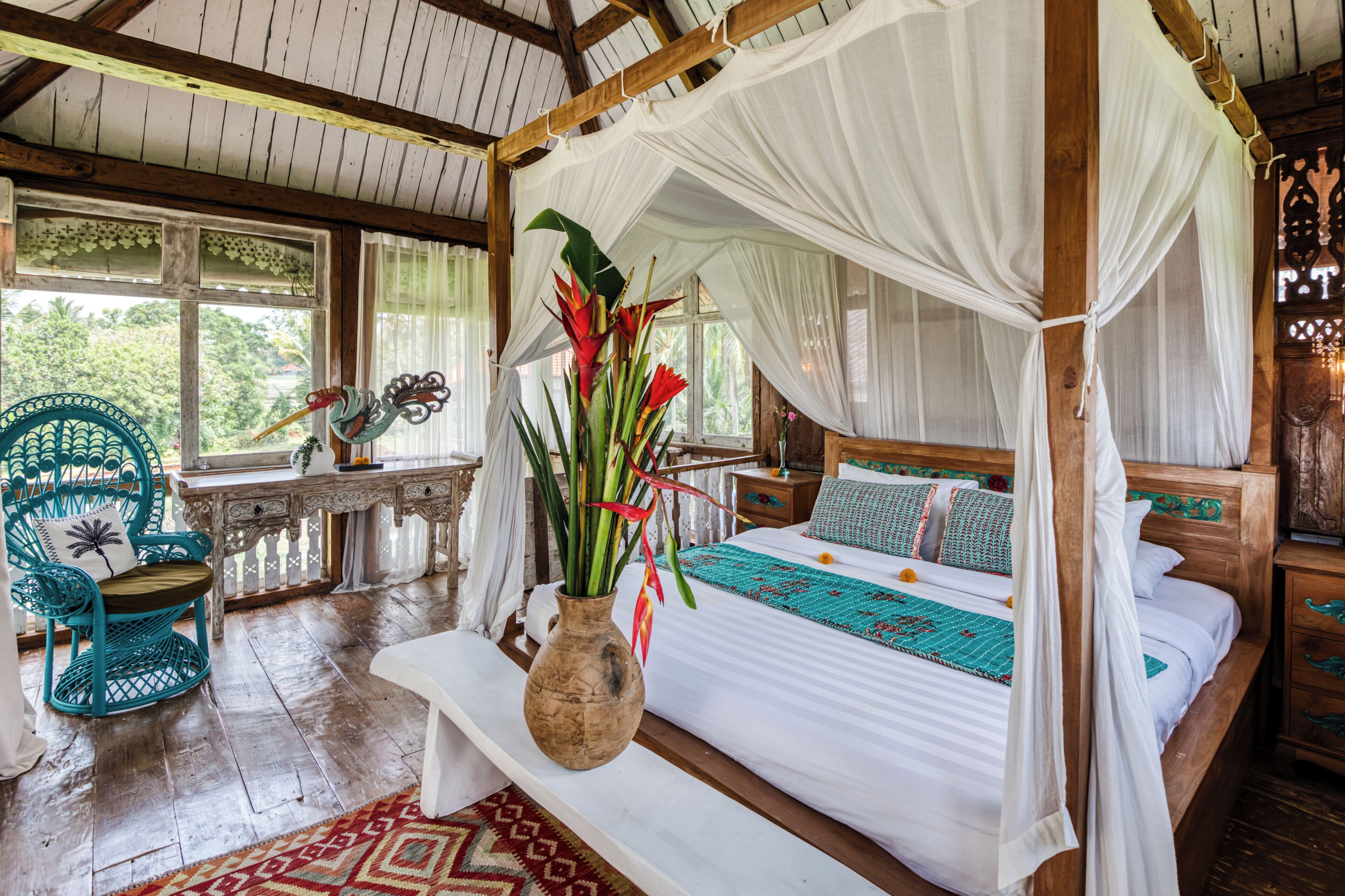 A room with a four-poster bed, a bench, a chair, a wooden desk, and a window with a view of greenery.