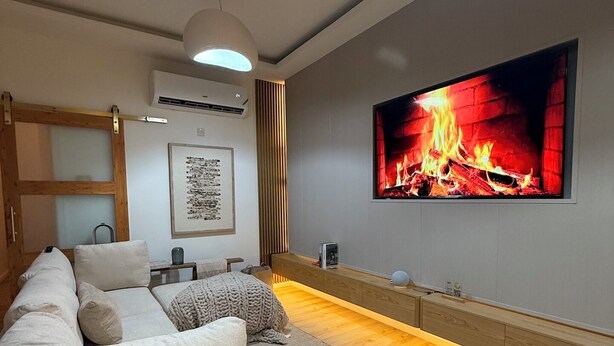 A modern living room with a large flat-screen TV displaying a fireplace, a white sofa, a wooden coffee table, and a small side table.