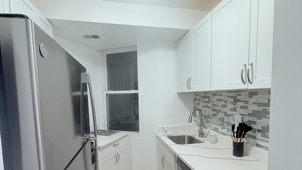 Una cocina compacta con armarios blancos, un refrigerador de acero inoxidable, salpicadero de azulejos y una ventana.
