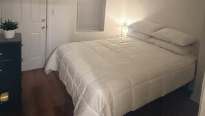 Un dormitorio con cama, cómoda, planta en maceta, ventana con persianas y luz de techo.