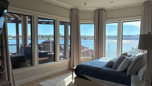 A bedroom with a large window overlooking a water body, a bed with a blue cover, and a ceiling fan.