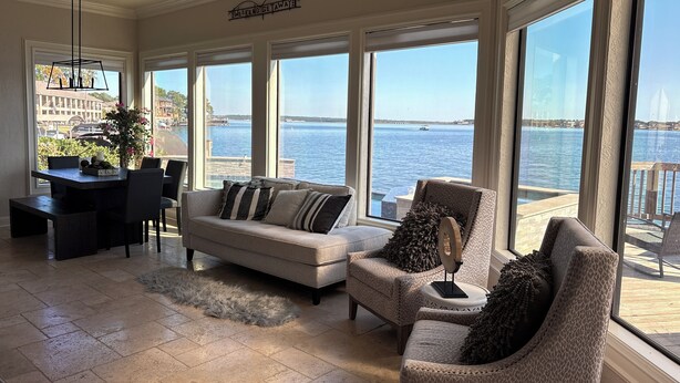 A spacious living room with a large window overlooking a body of water, a dining area with a table and chairs, and a cozy seating area with patterned cushions.