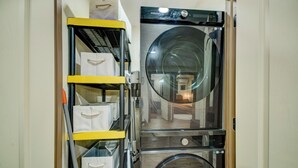 Ein kleiner Waschraum mit einer Frontlader-Waschmaschine und einem Trockner, einem gelben Wagen mit Aufbewahrungsbehältern und einem kleinen Spiegel an der Trockentür.