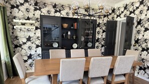 Comedor con mesa de madera y seis sillas, papel tapiz blanco y negro con motivos florales, vitrina con estantes y candelabro.