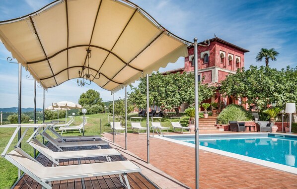 Un espace aménagé au bord de la piscine, avec des chaises longues, un auvent et un bâtiment rouge en arrière-plan.