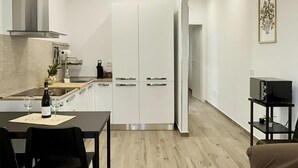 Et kompakt køkken med hvide skabe, et spisebord med stole, en kaffemaskine og et fjernsyn på et stativ.