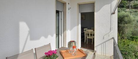 Een balkon met een houten tafel, stoelen, een rieten mand met bloemen en een dienblad met een glazen pot en een fles.