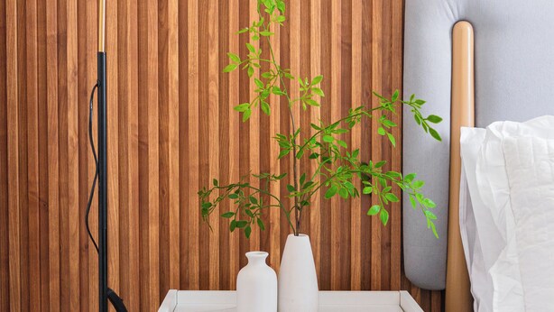 Een netjes ingerichte slaapkamer met een wand van houten latten, een nachtkastje met een witte vaas en een groene plant, een moderne vloerlamp en een bed met witte beddengoed.