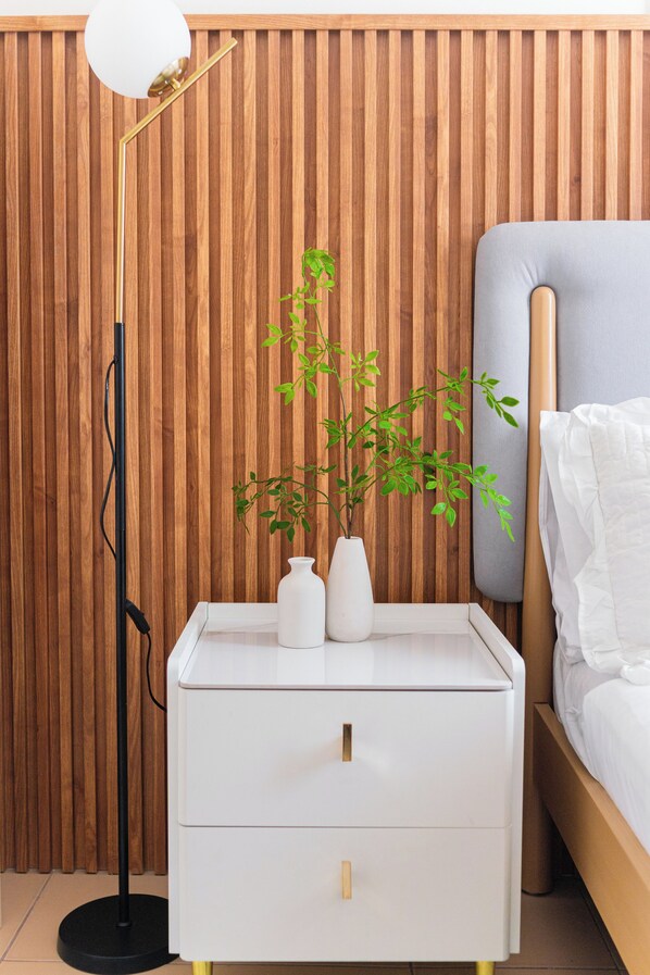 Un dormitorio ordenado con un cabecero de listones de madera, una mesita de noche con un jarrón blanco y una planta verde, una lámpara de pie moderna y una cama con ropa de cama blanca.